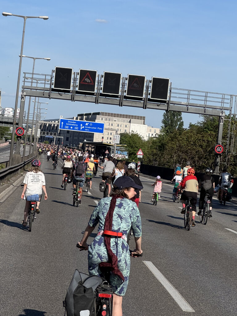 Das Bild zeigt eine große Gruppe von Fahrradfahrern, die auf einer mehrspurigen Autobahn fahren. Die Radfahrer scheinen an einer organisierten Veranstaltung teilzunehmen, denn sie fahren dicht beieinander und in einer Gruppe. Der Himmel ist klar und blau, und die Sonne scheint. Die Szene wirkt friedlich und fröhlich. Im Hintergrund sind Verkehrsschilder und Gebäude zu sehen. Ein Schild weist auf den Innsbrucker Platz hin. Einige der Radfahrer tragen Helme. Auf dem T-Shirt eines Radfahrers steht: „Sonne & Freunde sind die Dinge, die mich glücklich machen“.